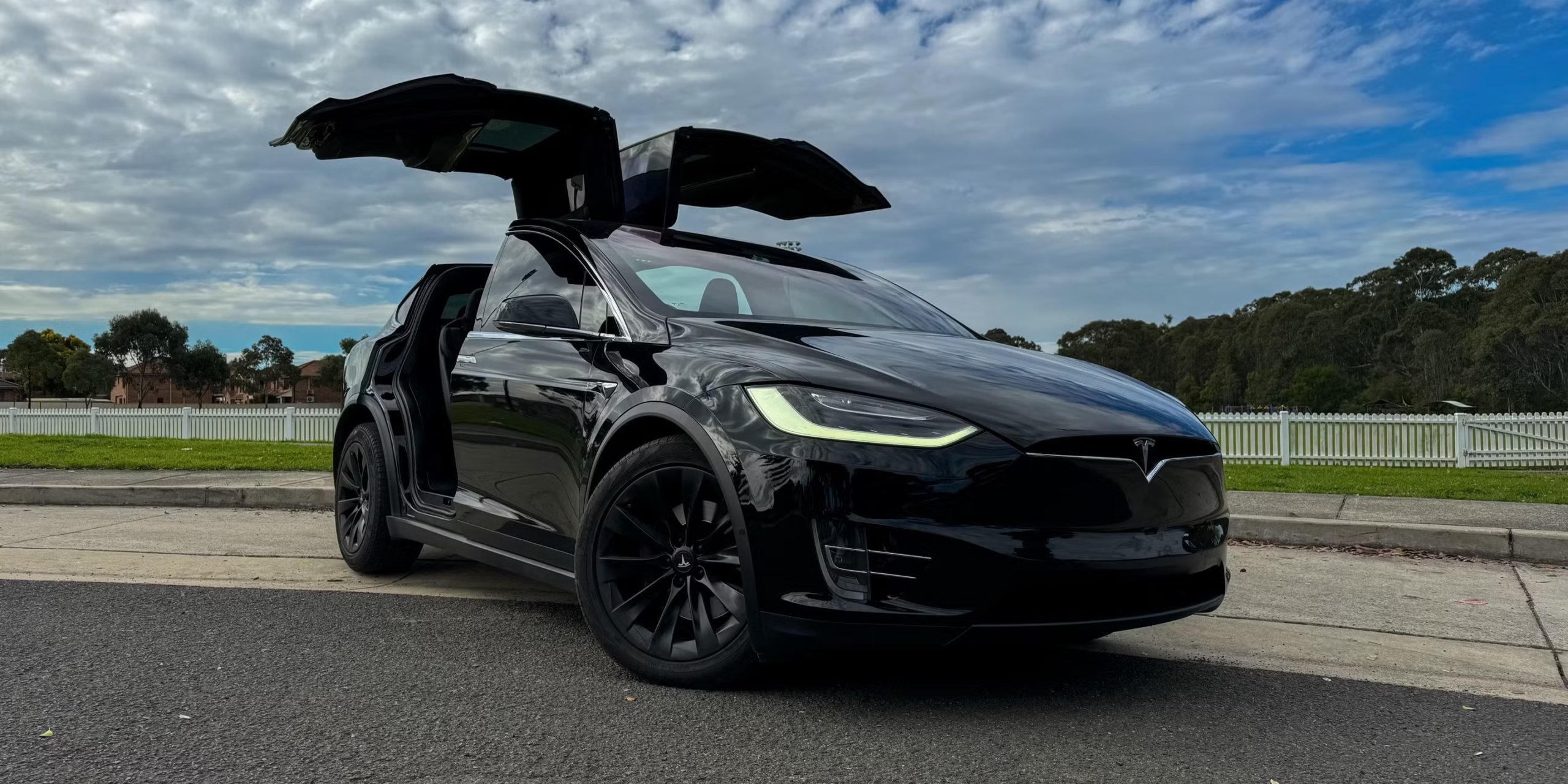 A black Tesla Model X with its falcon-wing doors open is parked on the street, with a white fence and trees in the background under a cloudy sky.